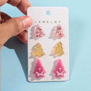 May include: A set of six earrings on a white card labeled "JEWELRY". The earrings include two pink glitter Christmas trees, two gold Christmas trees, and two pink glitter gnome shapes. The card is held by a hand against a blue background.