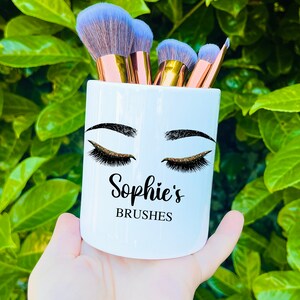 May include: White ceramic makeup brush holder with a black and gold eye design and the text "Sophie's Brushes".