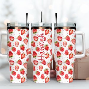 May include: Three stainless steel tumblers with white lids and handles. The tumblers are decorated with a pattern of red strawberries on a white background. The middle tumbler has the name "Hannah" written in red on the side.