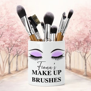 Könnte beinhalten: Weißer Keramik-Make-up-Pinselhalter mit einer stilisierten Illustration von Frauenaugen mit lila Lidschatten und langen Wimpern. Der Text "Fiona's MAKE UP BRUSHES" ist auf den Halter gedruckt.