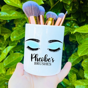 Könnte beinhalten: Weißer Keramik-Make-up-Pinselhalter mit einem blauen und schwarzen Augenmotiv und dem Text "Pheobe's Brushes".