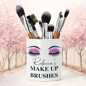 Puede incluir: Un portalápices de maquillaje de cerámica blanca con el texto "Rebecca's MAKE UP BRUSHES" en negro. El portalápices presenta un diseño de ojos cerrados con sombra de ojos de purpurina morada y largas pestañas negras. Varios pinceles de maquillaje están dentro.