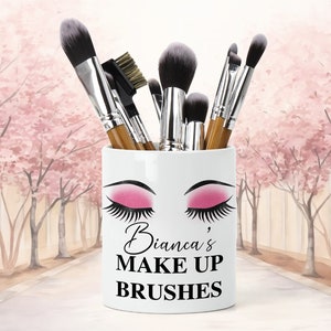 Puede incluir: Portacepillos de maquillaje de cerámica blanca con un diseño de ojo estilizado y sombra de ojos rosa. El portacepillos tiene el texto "Bianca's Make Up Brushes" impreso en él.