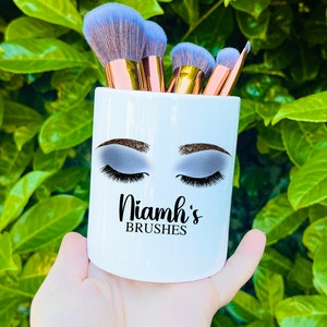 May include: White ceramic makeup brush holder with a stylized eye design and the text "Niamh's Brushes".