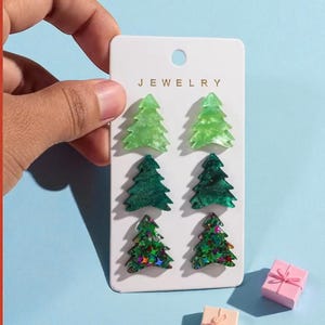 May include: A set of six Christmas tree-shaped earrings in varying shades of green, displayed on a white card labeled "JEWELRY". Two small gift boxes are also present. The earrings are made of a translucent material with glitter accents.