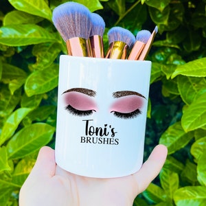 May include: White ceramic makeup brush holder with a stylized illustration of a woman's eyes and eyelashes. The holder has the text "Toni's Brushes" printed on the front.
