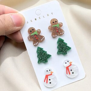 May include: A set of Christmas-themed stud earrings. The earrings include gingerbread men, green Christmas trees, and snowmen with red scarves. The earrings are on a white card with the word "JEWELRY" printed at the top.