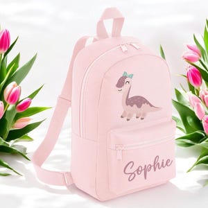 May include: Pink children's backpack featuring a cartoon dinosaur with a bow. The backpack has a zipped pocket and the name "Sophie" is embroidered on the front.