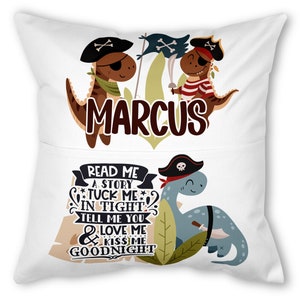 Op de afbeelding: Een wit kussen met een zakje aan de voorkant. Het kussen heeft een cartoon dinosaurus die een piratenhoed draagt en een zwaard vasthoudt. De tekst "MARCUS" is boven de dinosaurus gedrukt. Het zakje heeft een cartoon dinosaurus die een piratenhoed draagt en een zwaard vasthoudt. De tekst "READ ME A STORY TUCK ME IN TIGHT TELL ME YOU LOVE ME & KISS ME GOODNIGHT" is op het zakje gedrukt.
