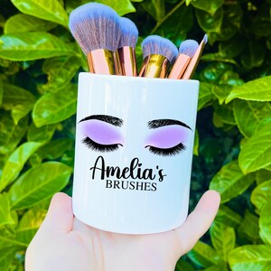 Könnte beinhalten: Weißer Keramik-Make-up-Pinselhalter mit einem Design aus geschlossenen Augen mit lilafarbenem Lidschatten und Wimpern. Der Halter trägt den Text "Amelia's Brushes".