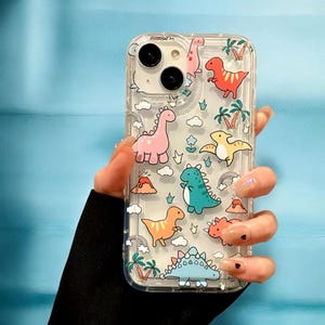Puede incluir: Carcasa transparente para teléfono con un diseño de dinosaurio colorido. La carcasa presenta una variedad de dinosaurios en diferentes colores, incluyendo rosa, verde, azul y naranja. La carcasa también presenta palmeras, nubes y volcanes.