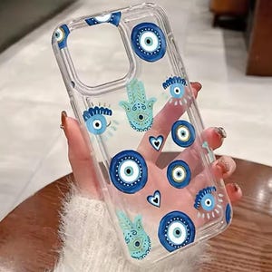 Könnte beinhalten: Transparente Handyhülle mit blauen und weißen Evil-Eye- und Hamsa-Hand-Motiven. Die Hülle zeigt verschiedene Augenformen, Herzen und Handsymbole. Die Hülle wird von einer Person gehalten.