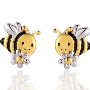 Könnte beinhalten: Ein Paar bienenförmige Ohrstecker. Die Ohrringe haben einen goldfarbenen Körper mit schwarzen Streifen, silbernen Flügeln und einer kleinen Blume. Die Bienen haben lächelnde Gesichter. Auf weißem Hintergrund.
