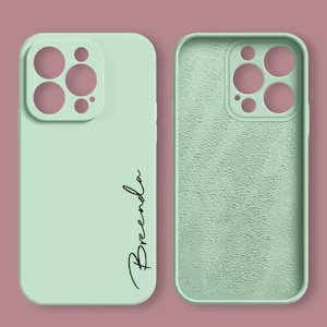 May include: A light green phone case with a textured back and a clear camera lens cutout. The front of the case has the name "Brenda" written in black cursive.