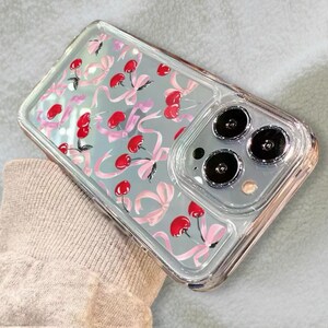 May include: A clear phone case with a design of pink bows and red cherries. The case has a raised camera bump with three black camera lenses.