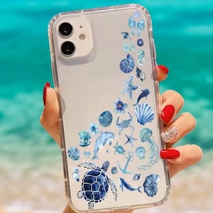Può includere: Una custodia per telefono trasparente con un design acquerellato blu e bianco di vita marina, tra cui una tartaruga, un delfino, una stella marina, conchiglie e un'ancora.
