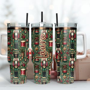 May include: Three stainless steel tumblers with a Christmas nutcracker pattern. The tumbler on the left has a white handle. The tumbler in the middle has the name "Laura" printed on it.