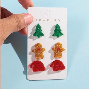 May include: A set of Christmas-themed stud earrings on a white card. The earrings include green Christmas trees, gingerbread men, and red Santa hats. The word "JEWELRY" is printed at the top of the card.