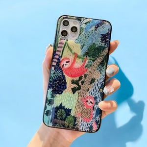 May include: A phone case with a colorful design featuring sloths, leaves, and gold glitter. The case has a clear background and a black border.
