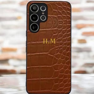 Peut inclure: Une coque de téléphone en cuir marron avec un motif crocodile et les initiales "H.M." en lettres dorées.
