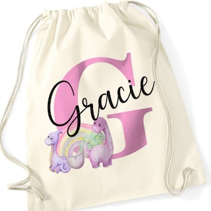 May include: A white canvas drawstring bag with a pink letter G and the name "Gracie" in black script. The bag features a pink dinosaur with a rainbow and a green palm leaf.