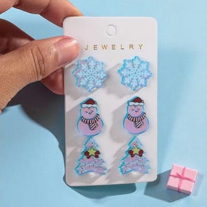 May include: A set of Christmas-themed stud earrings on a white card. The earrings feature snowflake, snowman, and Christmas tree designs in shades of blue, pink, and red. A small pink gift box with a pink ribbon is also present.