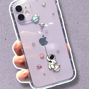 Könnte beinhalten: Transparente Handyhülle mit lila Rückseite mit einem Cartoon-Astronauten, Planeten und Sternen. Der Astronaut hält eine Schnur, die an einem Planeten befestigt ist. Die Hülle hat einen transparenten Stoßfänger.