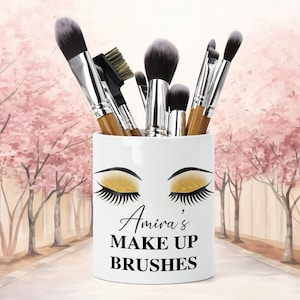 Könnte beinhalten: Weißer Keramik-Make-up-Pinselhalter mit einem goldenen Glitzer-Augen-Design und dem Text "Amira's Make Up Brushes".