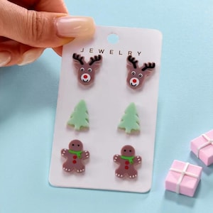 May include: A set of Christmas-themed stud earrings on a white card. The earrings feature reindeer, green Christmas trees, and gingerbread men. The word "JEWELRY" is printed at the top of the card. The background is light blue.