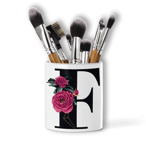 Puede incluir: Portacepillos de maquillaje de cerámica blanca con una letra "F" negra y rosas rosas. El portacepillos está lleno de pinceles de maquillaje.