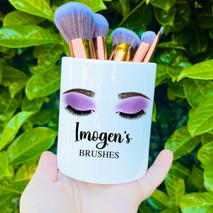 Könnte beinhalten: Weißer Keramik-Make-up-Pinselhalter mit einem Design aus einem Paar Augen mit lila Lidschatten und Wimpern. Der Text "Imogen's Brushes" ist unter den Augen aufgedruckt.