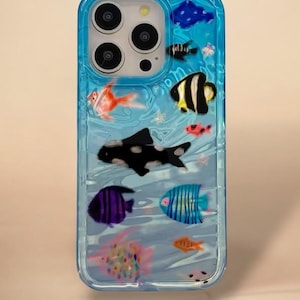 Könnte beinhalten: Eine transparente Handyhülle mit blauem Rand, die eine lebendige Unterwasserszene zeigt. Bunte Fische in verschiedenen Formen und Größen, darunter orange, schwarz-weiß und lila, schwimmen vor einem hellblauen Hintergrund. Die Hülle hat ein glänzendes Finish.