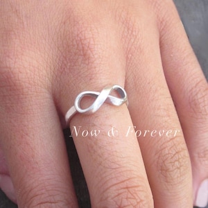 Puede incluir: Un anillo de plata con forma de infinito se lleva en un dedo. El anillo es un bucle continuo, que simboliza la eternidad. Las palabras "Now & Forever" están impresas en el dedo.