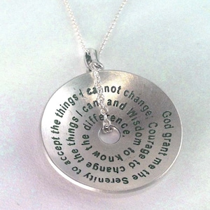 Puede incluir: Un collar con colgante plateado con un dije circular. El dije presenta texto grabado, incluidas las palabras "Serenidad", "Coraje" y "Sabiduría". El collar tiene una cadena delicada.