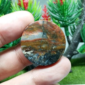 Puede incluir: Una piedra pulida y redonda con una paleta de colores natural y terrosa. La piedra presenta una mezcla de tonos marrones, rojos, blancos y negros, con grietas y patrones visibles. La piedra se sostiene en una mano, mostrando su tamaño y textura.