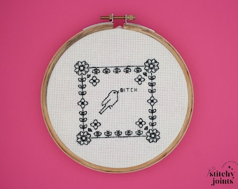 May include: A cross-stitch embroidery hoop with a black and white design. The design features a bird, floral border, and the word "BITCH". The embroidery is set against a white fabric and framed by a wooden hoop, set against a pink background.