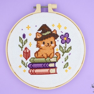 Könnte beinhalten: Eine Kreuzstichstickerei in einem runden Holzrahmen mit einer orangefarbenen Katze, die einen braunen Hexenhut trägt und auf einem Stapel aus drei Büchern sitzt. Das Motiv enthält eine violette Blume, einen roten Kristall und gelbe Sterne.