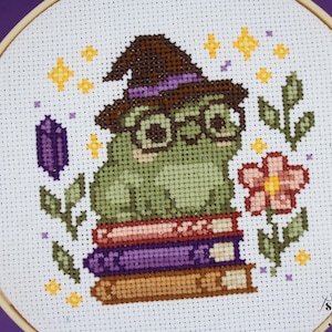 May include: Cross-stitch embroidery of a green frog wearing a witch hat and glasses, sitting on a stack of books. The design includes a purple crystal, a pink flower, and yellow stars, all within a wooden hoop.