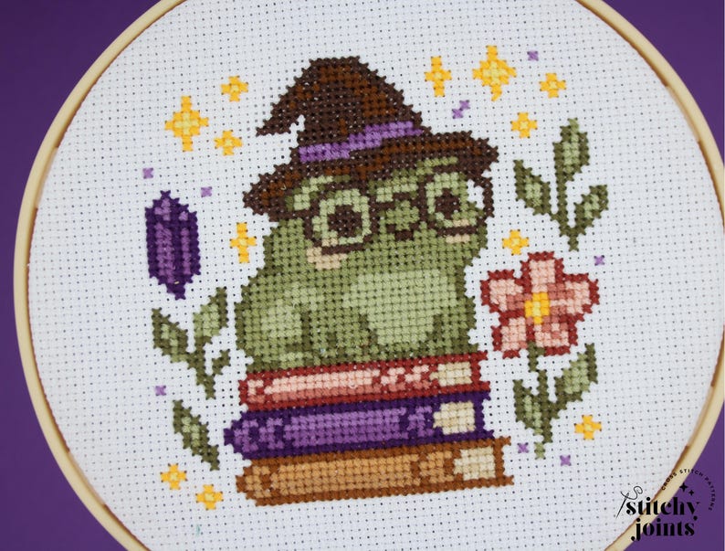 May include: Cross-stitch embroidery of a green frog wearing a witch hat and glasses, sitting on a stack of books. The design includes a purple crystal, a pink flower, and yellow stars, all within a wooden hoop.