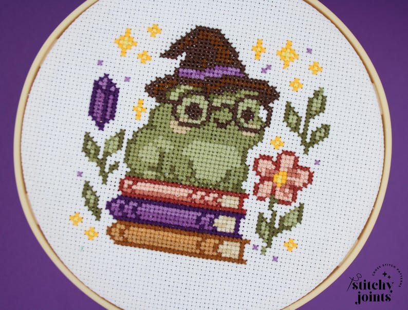 May include: Cross-stitch embroidery of a green frog wearing glasses and a witch hat, sitting on a stack of books. The design includes a purple crystal, a pink flower, and yellow stars, all within a wooden embroidery hoop.