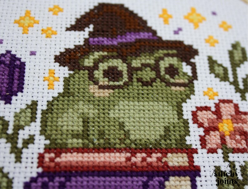 May include: Cross-stitch art featuring a green frog wearing a witch hat and glasses. The frog is sitting on a purple book. The background includes flowers, stars, and foliage. The color palette includes green, brown, purple, and yellow.