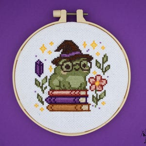 Może przedstawiać: Haft krzyżykowy przedstawiający zieloną żabę w kapeluszu czarownicy i okularach, siedzącą na stosie książek. Projekt zawiera kwiat, fioletowy kryształ i gwiazdy, wszystko w drewnianym tamborku.