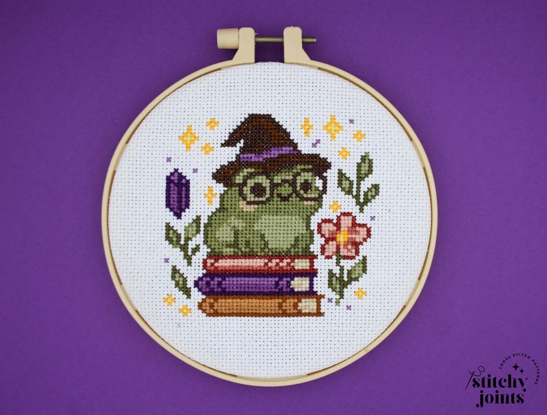 May include: Cross-stitch embroidery of a green frog wearing a witch hat and glasses, sitting on a stack of books. The design includes a flower, a purple crystal, and stars, all within a wooden embroidery hoop.