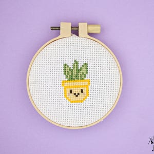 Puede incluir: Un bastidor de bordado de punto de cruz con un cactus sonriente en maceta amarilla con hojas verdes. El bordado está sobre una tela blanca y enmarcado por un aro de madera. El fondo es de color lavanda.