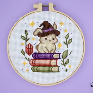 Può includere: Ricamo a punto croce in un telaio di legno. Il disegno presenta un coniglietto con un cappello da strega, seduto su una pila di libri. Il disegno include un cristallo rosso, stelle e fogliame verde. Lo sfondo è bianco.