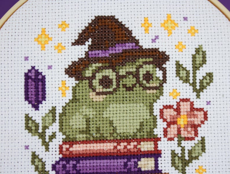 May include: Cross-stitch embroidery of a green frog wearing glasses and a witch hat, sitting on books. The design includes a flower, leaves, stars, and a purple crystal, all on a white background.