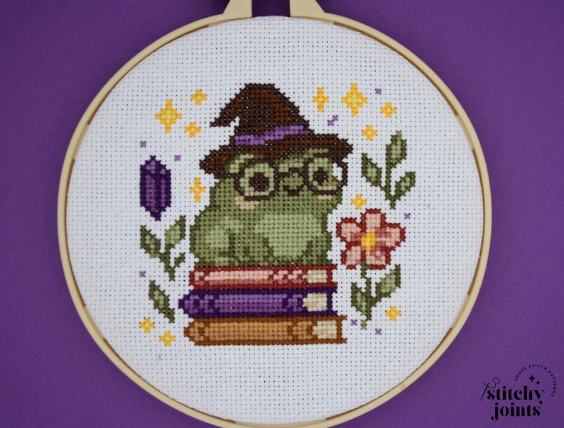 May include: Cross-stitch embroidery in a wooden hoop features a green frog wearing a witch hat and glasses, sitting on a stack of books. The design includes a purple crystal, stars, leaves, and a pink flower. The background is white.