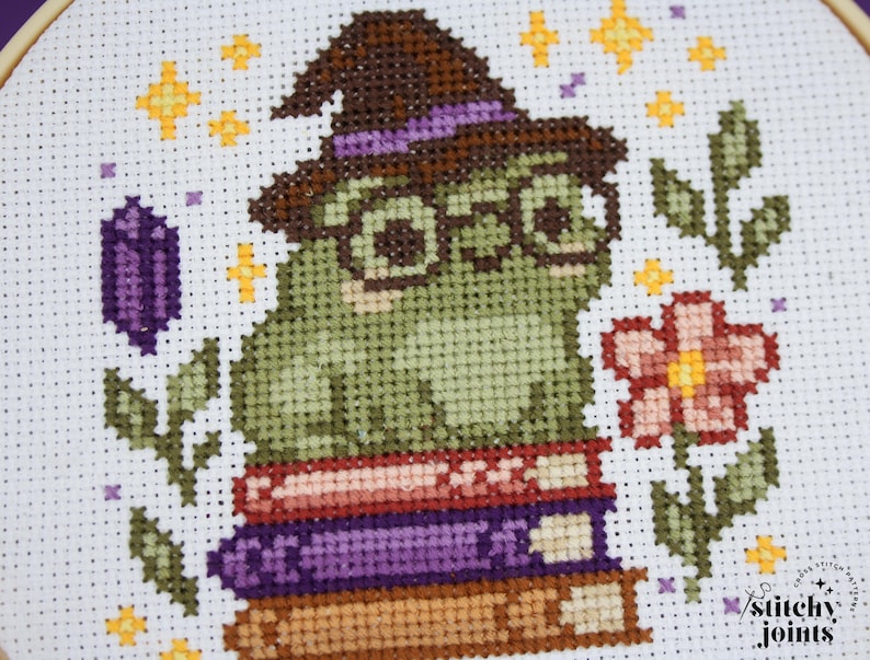 May include: Cross-stitch embroidery of a green frog wearing a witch hat and glasses, sitting on a stack of books. The design includes a purple crystal, flowers, and stars. The text "stitchy joints" is in the lower right corner.