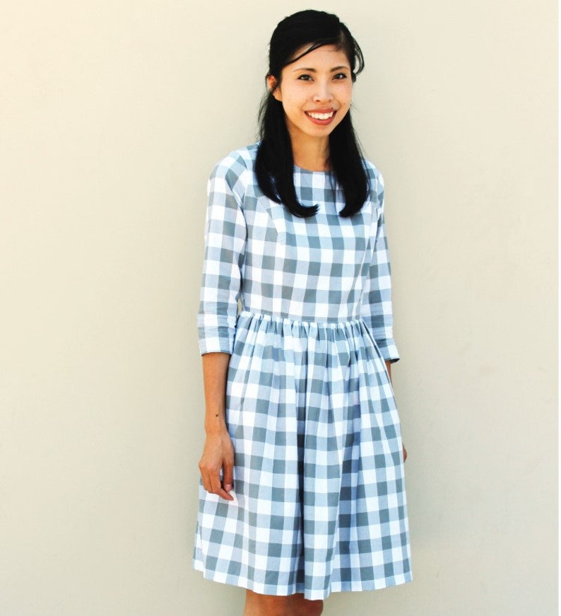 May include: A woman wearing a gray and white gingham dress with 3/4 sleeves. The dress has a fitted bodice and a full skirt.