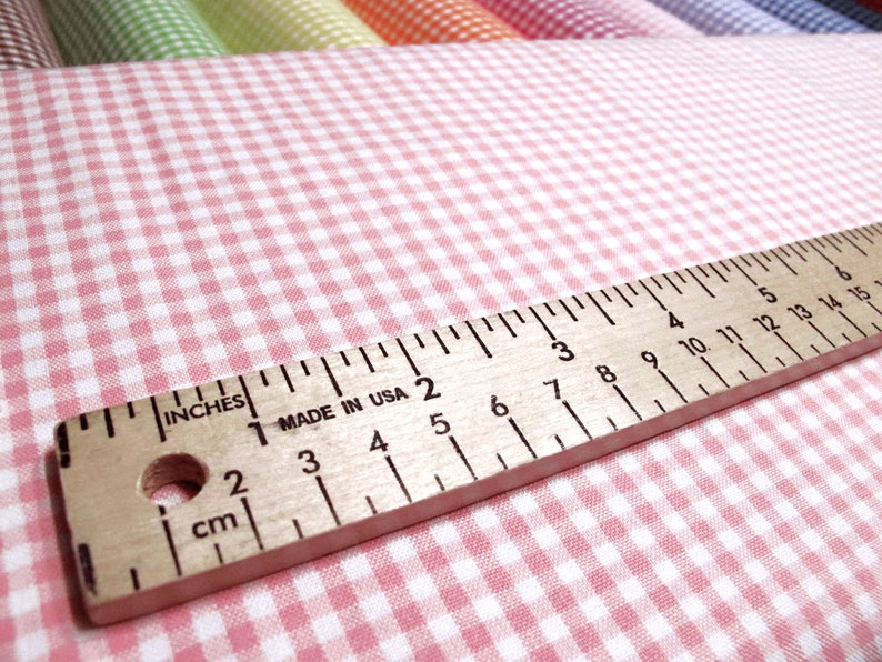May include: A light pink and white gingham fabric featuring a small checkered pattern. A wooden ruler is placed across the fabric, showing markings in inches and centimeters, with the text "INCHES", "MADE IN USA", and "cm" printed on it.